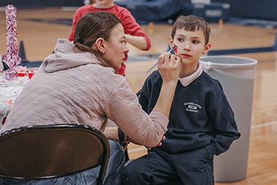 A student getting face painted by a parent at a Valentine's Day party. 